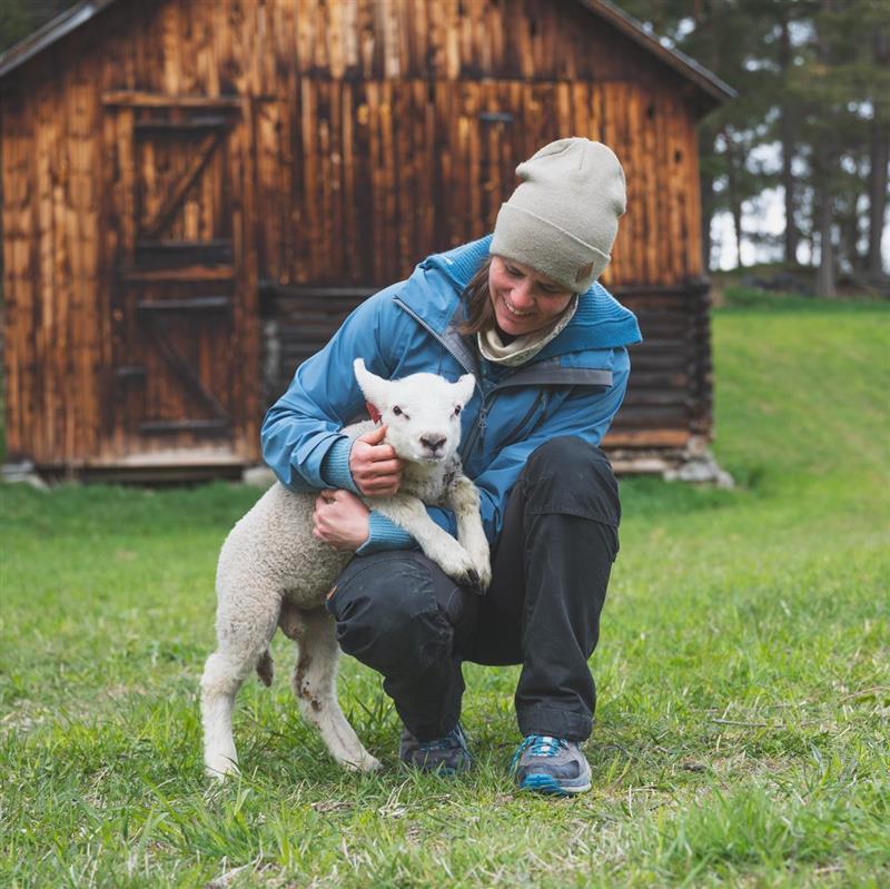 Kvinnelig bonde med lam foran låve